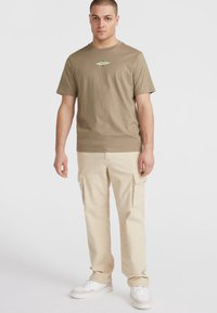 Khaki t-shirt med et centralt grønt grafik, parret med beige cargo bukser med sidelommer og hvide sneakers.