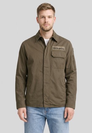 Man draagt een olijfgroene blouse met knopen, borst- en armzakken en een "Blauer:" logo patch, gecombineerd met lichtblauwe spijkerbroek.