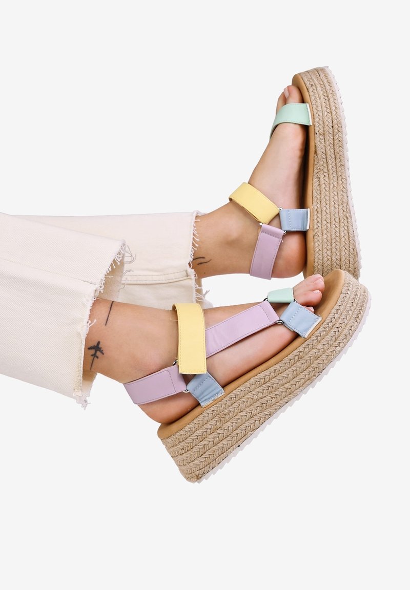 Pieds portant des sandales plateforme à blocs de couleurs pastel avec semelles en corde tissée et pantalons beige clair à ourlet effiloché sur fond blanc.