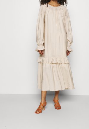 Beige lange jurk met lange mouwen, ruches aan de manchetten en zoom, gemaakt van gestructureerde stof. Gecombineerd met oranje sandalen met hoge hak en bandjes.