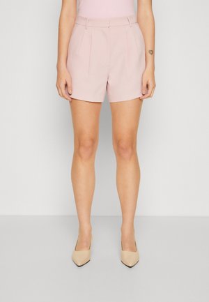 Mujer con pantalones cortos rosa claro hechos a medida con pliegues, de pie recta, combinada con tacones beige de punta afilada sobre un fondo liso.