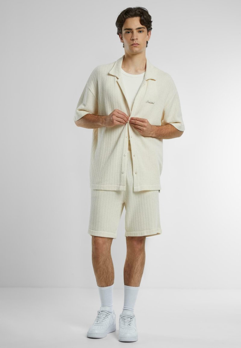 Kremfarget strikket kortermet skjorte og matchende shorts. Har et teksturert mønster, knappeåpning og minimal merkevare.