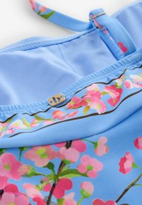 Primer plano de tela azul claro con estampado floral rosa y un pequeño detalle de logo metálico en forma de "D" en una correa con un anillo metálico.