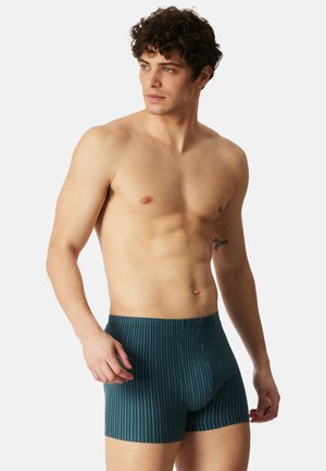 Alaston ylävartalo, kiharat hiukset, tummaa vihreää bokserialushousuja, joissa ohuet valkoiset pystysuorat raidat, katsoo sivulle yksinkertaisen taustan edessä.