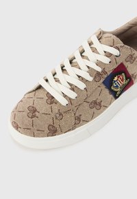Beige mönstrad sneakers av kanvas, med vita snörningar, en vit gummisula och en röd, blå och guld emblem patch på sidan.