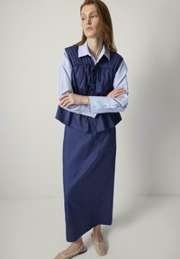 En modell står med armarna i kors, klädd i en marinblå lagerklänning över en ljusblå skjorta och bruna slip-on skor, mot en neutral bakgrund.