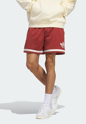 Persona que lleva shorts de malla rojos de Adidas con logo y ribete blancos, sudadera con capucha crema, calcetines blancos y zapatillas Adidas blancas, de pie con las piernas cruzadas.