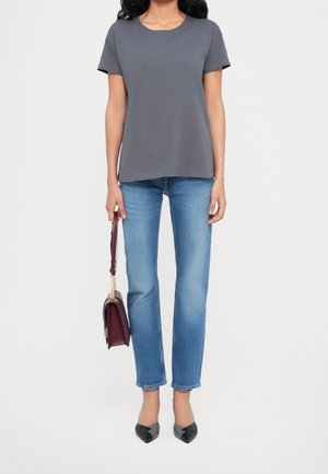 Vrouw met een grijs topje met korte mouwen, lichtblauwe spijkerbroek, zwarte puntige schoenen en een bordeauxrode handtas met een kettingband.