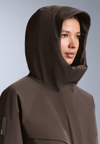 Hnedá nepremokavá bunda s vysokým golierom a nastaviteľnou kapucňou, s hladkým povrchom a prednými vreckami na hrudi s klopami.