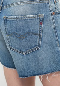 Τζιν σορτς σε ανοικτό μπλε χρώμα με ξεφτισμένο τελείωμα. Η πίσω τσέπη διαθέτει περίπλοκο σχέδιο ραφής και μικρή κόκκινη λεπτομέρεια λογότυπου.
