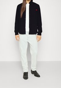 Camisa negra de botones con un pequeño logo rojo en el pecho, combinada con jeans azul claro y botas negras de tobillo. Tela suave y ajuste estándar.