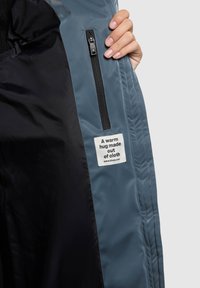 Sinisen takin sisäpinta, jossa on sileä pinta, vasemmalla vetoketjullinen tasku ja etiketti, jossa lukee "Lämmin halaus kankaasta."