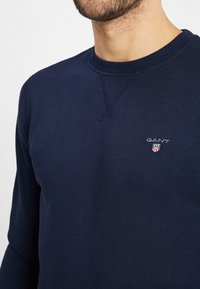 Man som bär en marinblå sweatshirt med rund halsringning och GANT-logo samt sköldemblem på bröstet, synlig från nacken till mitten av överkroppen.