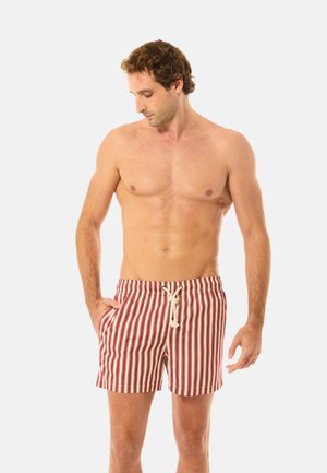 Herrenloser Mann mit lockigem Haar, trägt rot-weiß gestreifte Schwimmshorts mit weißem Kordelzug, Hände in den Taschen, schaut nach unten links.