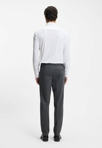 Man die met zijn rug naar voren staat, draagt een wit overhemd met lange mouwen, grijze pantalon en zwarte nette schoenen, tegen een effen witte achtergrond.