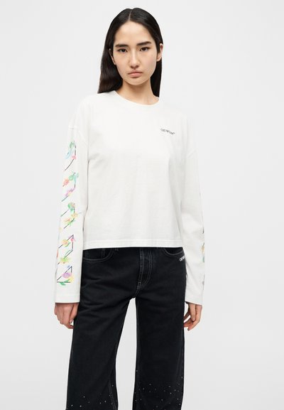 Λευκό πουκάμισο με μακριά μανίκια και λουλουδένια σχέδια στα μανίκια, με κοντό κόψιμο και διακριτικό λογότυπο στο στήθος. Συνδυασμένο με μαύρο τζιν.