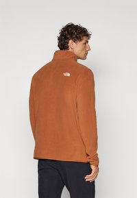 Veste en polaire couleur rouille avec un col haut. Elle présente un petit logo blanc à l'arrière, une texture lisse et un design décontracté.