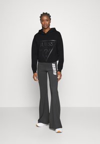 Juodas trumpas hoodie su tekstūruotu logotipu ir virvele, derinamas su pilkomis platėjančiomis kelnėmis, turinčiomis baltą logotipą vienoje pusėje ir baltais batais.