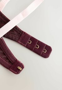 Nærbillede af en bordeaux bh-strop med hægte- og øjnelukning og en lys pink justerbar skulderstrop på en lys baggrund.