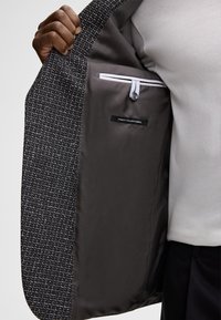 Schwarzer und weißer Textilstoff Blazer-Innenraum, der einen glatten grauen Lining zeigt, mit einem Etikett mit der Aufschrift "FINEST ITALIAN FABRIC" und einer Tasche mit Knopfverschluss.
