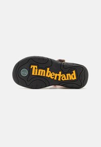 Svart gummisåle med et fremtredende gult "Timberland"-logo, teksturerte mønstre for grep, og et rundt logo på hælen.