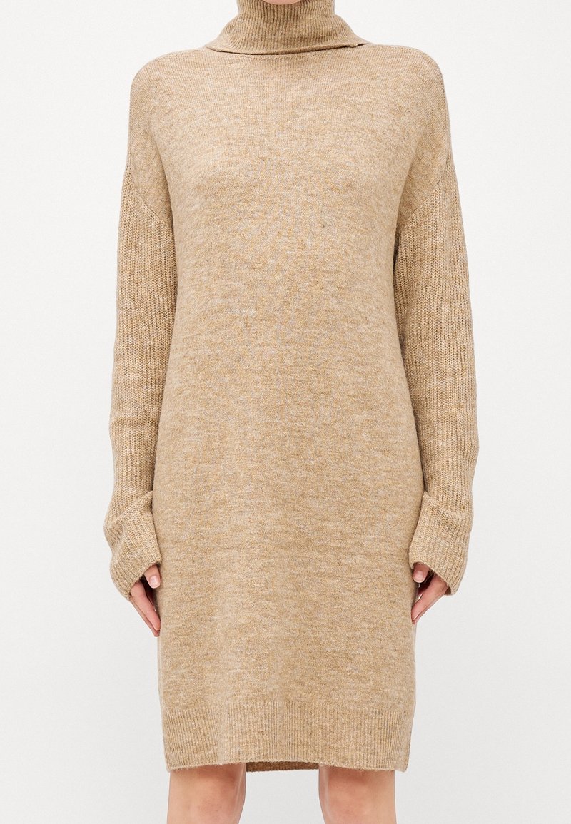 Robe-pull beige en maille avec col roulé, manches longues, poignets, ourlet et col côtelés, portée par une personne debout.