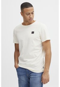 Cremefarvet t-shirt i bomuld med rund hals og korte ærmer. Inkluderer et sort logo-patch på brystet. Båret med blå jeans.