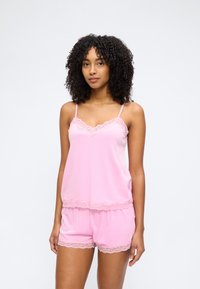 Γυναίκα με σγουρά μαλλιά, που φοράει ροζ καμισόλι με δαντέλα και αντίστοιχα σορτς, στέκεται μπροστά σε απλό ανοιχτό φόντο.