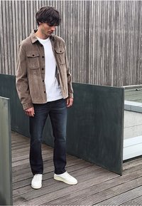 Mladý muž oblečený v bílém tričku, hnědé bundě, tmavých džínách a bílých teniskách stojí na dřevěné terase vedle moderního plota.