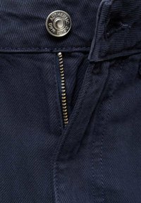 Mörkblå denimbyxor har en silverknapp med "AUTHENTIC" inristad, en guldfärgad dragkedja och textil med synliga stygn.