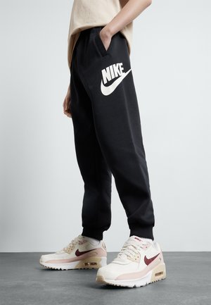 Person iført sorte Nike træningsbukser med stort hvidt logo og hvide Nike sneakers med mørkerødt swoosh, der står på gråt gulv.