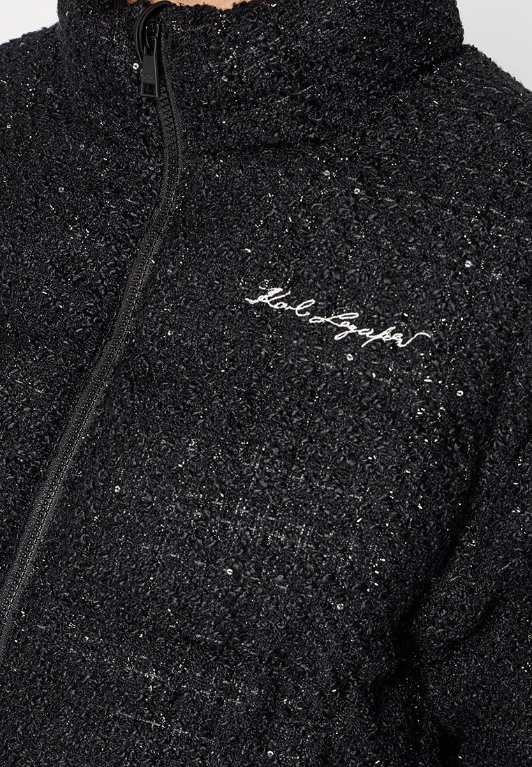 Schwarze, strukturierte Jacke mit Reißverschluss, ausgestattet mit einem plüschigen Stoff und silbernen Akzenten. Mit einem weißen Logo auf der Vorderseite.
