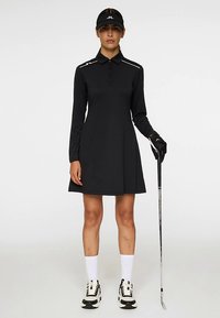 Schwarzes Golfkleid mit langen Ärmeln, Knopfleiste, weißen Akzentstreifen auf den Schultern, kombiniert mit schwarzem Visor, Handschuh und Golfschläger.