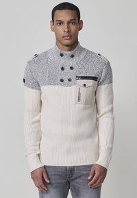 Suéter de punto para hombre con un diseño de bloques de color gris y crema, cuello alto, detalles de botones y un bolsillo en el pecho con cremallera.