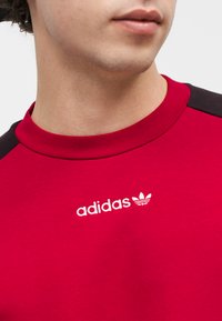 Άνδρας που φοράει κόκκινο μπλουζάκι Adidas με μαύρες λεπτομέρειες στους ώμους και λευκό λογότυπο Adidas στο στήθος, ορατός λαιμός.