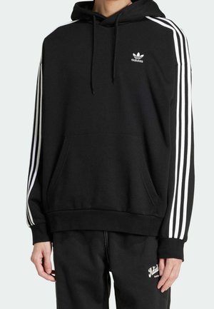 Sweat à capuche Adidas noir avec trois bandes blanches sur les manches, poche avant et logo sur la poitrine gauche, porté avec un pantalon noir assorti.