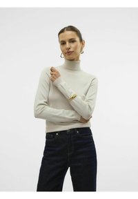 Maglione beige a collo alto con design aderente, maniche lunghe e collo a coste; indossato con jeans blu scuro. Bracciale dorato al polso.