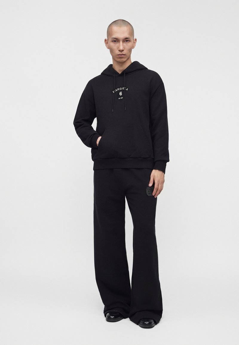 Homme debout portant un sweat à capuche noir avec un logo sur le devant, un pantalon noir ample et des chaussures noires sur un fond gris uni.