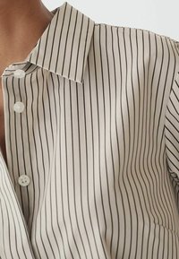 Gros plan sur une chemise de couleur crème avec de fines rayures verticales noires et des boutons blancs sur la poitrine et l'épaule d'une personne.