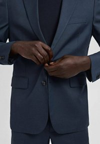 Czarnoskóry mężczyzna zapinający granatowy żakiet na czarną koszulę, widoczny od połowy torsu do rąk.