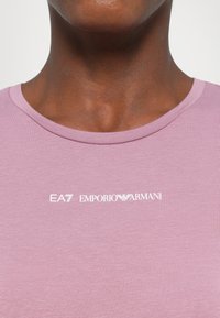 Rožinė medvilninė marškinė su apvaliame kakle, ant krūtinės puikuojasi baltas "EA7 EMPORIO ARMANI" logotipas. Lygi tekstūra ir prigludusi konstrukcija.