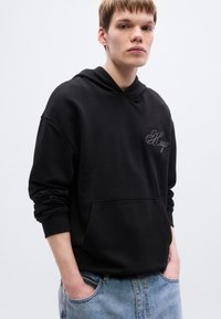 Μαύρο φούτερ με κουκούλα από βαμβάκι, με μπροστινή τσέπη και κεντημένο λογότυπο. Το μοντέλο φοράει τζιν παντελόνι σε ανοιχτό μπλε χρώμα.