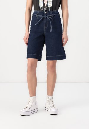 Person, der bærer mørkeblå højtaljede denimshorts til knæet med bundet bælte i taljen og hvide høje sneakers.