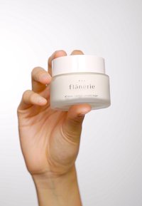 Et hvitt, rundt glass med et lys rosa lokk, merket "flânerie," som inneholder 45 ml med vegansk krem, holdt av en hånd mot en uskarp bakgrunn.