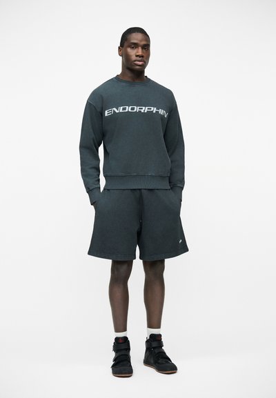 Jeune homme debout, les mains dans les poches, portant un sweat-shirt gris foncé "ENDORPHIN", un short assorti, des chaussettes blanches et des baskets montantes noires.