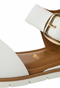 Sandales blanches avec une large sangle et un détail de boucle dorée, semelle intérieure marron et semelle blanche. Texture lisse avec un design simple et moderne.