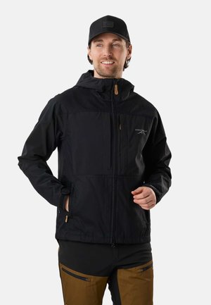 Man wearing black hooded outdoor jacket, black cap, and brown pants with black zipper pockets, smiling and looking right.