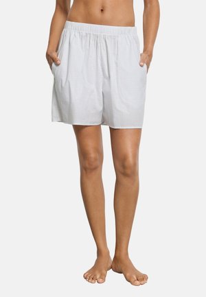 Unbedeckter Mittelteil und Beine einer Person, die lockere, weiße Shorts mit elastischem Bund trägt, mit den Händen in den Taschen steht und barfuß vor einem schlichten Hintergrund steht.