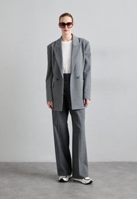 Blazer gris ajusté à double boutonnage sur un t-shirt blanc, associé à un pantalon gris à jambes larges et des baskets blanches. Grandes lunettes de soleil sombres.