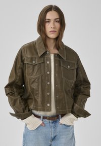 Tânără cu păr drept, castaniu, purtând o jachetă din piele verde măsliniu, pulover bej, blugi albaștri și mănuși fără degete, stând cu mâinile în buzunare.